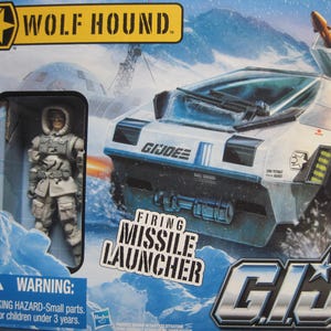 May include: A white toy military vehicle with a missile launcher and the words "Firing Missile Launcher" printed on the side. The vehicle is labeled "GI Joe" and "Wolf Hound". A toy soldier is standing in the background.