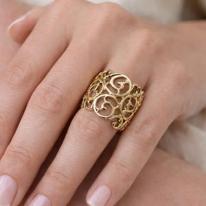 Può includere: Un anello color oro con un design traforato con motivi a spirale. L'anello è largo e si trova su un dito, mostrando i suoi intricati dettagli e la calda tonalità metallica. Un accessorio di gioielleria.