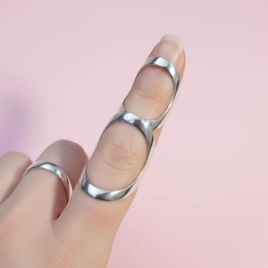May include: A silver ring and a silver finger ring are displayed against a pink background. The finger ring has two connected loops that cover two finger segments. The other ring is a simple band.