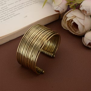 Può includere: Un bracciale a polsino color oro con più bande parallele. Il design aperto ne facilita l'indossatura. Il bracciale è in metallo e ha un aspetto vintage. Il gioiello è esposto su una superficie marrone.