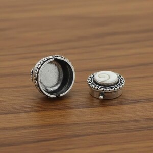May include: Two small, silver-colored trinket boxes on a wooden surface. One box is open, revealing the interior. The other box is closed, with a white, shell-like design on the lid. The boxes have ornate, beaded edges.
