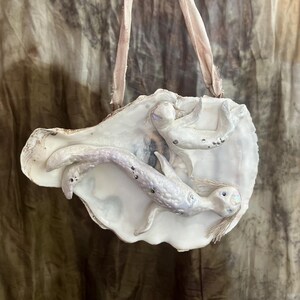 Könnte beinhalten: Ein dekoratives Austernschalen-Ornament mit zwei modellierten, schillernd weißen Meerjungfrauenfiguren auf der Oberfläche. Die Schale ist an einem ausgefransten, hellrosa Stoffband befestigt und bildet ein hängendes Objekt mit Perlmuttglanz.