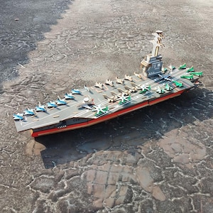 May include: A wooden model of an aircraft carrier with numerous miniature airplanes on the deck. The carrier is painted gray, red, and brown, with a tower structure and the number 78 visible. The model is set on a textured surface.