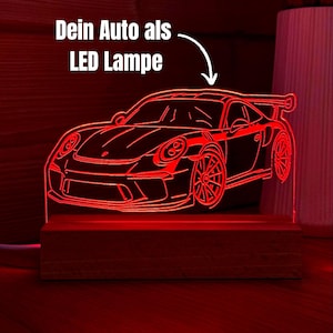 Könnte beinhalten: Eine rote LED-Lampe mit einer transparenten Acrylplatte, die den Umriss eines Sportwagens zeigt. Die Lampe ist auf einem rechteckigen Holzsockel montiert. Über dem Auto steht der Text "Dein Auto als LED Lampe" mit einem weißen Pfeil.