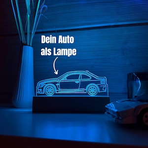Könnte beinhalten: Eine blaue LED-Lampe in Form eines Autos mit dem Text "Dein Auto als Lampe". Die Lampe ist auf einem dunklen Holzsockel montiert. Links ein weißes Gefäß mit grünen Stielen, rechts ein Spielzeugauto.