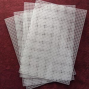 May include: A stack of five rectangular white plastic canvas sheets, each featuring a uniform grid of square holes. The sheets are arranged in a fanned-out pattern, showcasing the rigid, perforated structure of the material used for needlepoint.