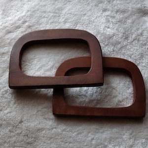 May include: Two dark brown wooden purse handles with a rectangular shape and rounded top. The handles are interlocked, showing their design and construction. The wood grain is visible, adding to the natural aesthetic.