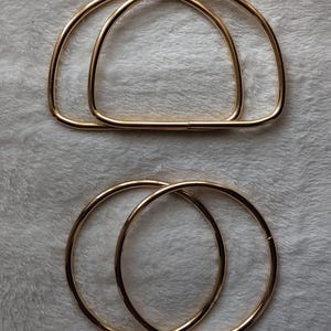 Op de afbeelding: Twee goudkleurige D-vormige tassenhandvatten van metaal en twee ronde tassenhandvatten van metaal. De D-vormige handvatten hebben een platte basis en een gebogen bovenkant, terwijl de ronde handvatten volledige ringen zijn. Gladde, gepolijste afwerking.