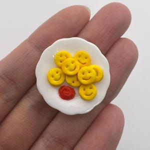 Può includere: Un piatto bianco in miniatura con pancake gialli a forma di faccina sorridente e una macchia rossa di ketchup. Il piatto è tenuto in mano da una persona su sfondo bianco. I pancake hanno sorrisi neri.