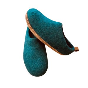 May include: A pair of teal felted wool slippers featuring a rounded toe design and a tan-coloured sole. The slippers have a textured, fuzzy surface and a small pull tab at the heel, showcasing a slip-on style with a deep opening for the foot.