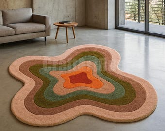 May include: A large, abstract rug with a unique, organic shape. The rug features concentric layers of color, including shades of peach, brown, green, and orange. The rug is placed on a concrete floor in a modern living space.