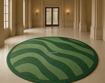 May include: An oval-shaped green rug with a wavy pattern in varying shades of green. The rug has a dark green border and is placed in a large, well-lit room. The text "CraftArt RUGS" is visible on the rug.