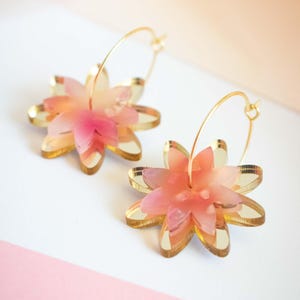 Puede incluir: Un par de pendientes con forma de flor con aros dorados. Los pendientes presentan pétalos en capas en tonos rosa y melocotón, con un contorno dorado. Se muestran sobre una superficie blanca y rosa.