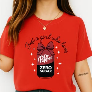 May include: Red t-shirt with the text "Just a girl who loves" above a graphic of a Dr. Pepper can with a bow. The can design includes the words "Dr. Pepper" and "Zero Sugar". White star shapes surround the can.