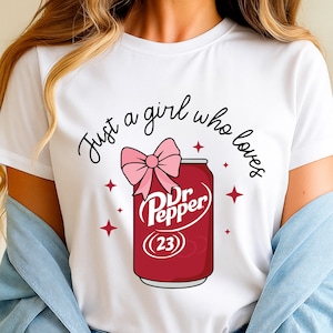 May include: White t-shirt with the text "Just a girl who loves" above a cartoon image of a Dr. Pepper can with a pink bow. The can is red with the Dr. Pepper logo and the number 23. Pink star accents surround the can.