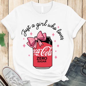 Könnte beinhalten: Weißes T-Shirt mit einem Coca-Cola Zero Sugar-Dosen-Design, einer rosa Schleife und dem Schriftzug "Just a girl who loves". Rosa Diamanten umgeben die Dose. Das Shirt wird mit blauen Jeans und weißen Turnschuhen kombiniert.