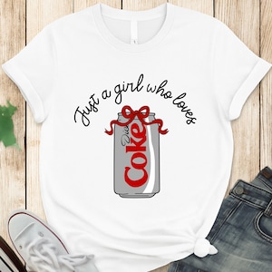 May include: White t-shirt featuring a Diet Coke can graphic with a red bow and the text "Just a girl who loves." The can is gray with red lettering. The shirt is paired with blue jeans and white sneakers.