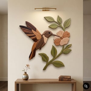 May include: A wall-mounted wooden art piece featuring a hummingbird in flight with brown and tan wood tones, approaching a light-coloured wooden flower on a green leafy stem. The sculpture is composed of layered wood segments with visible grain.