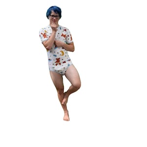May include: A white onesie featuring a pattern of brown teddy bears, yellow moons, and blue clouds. The onesie has short sleeves and a matching bottom. The person is wearing glasses and has blue hair.