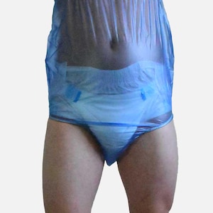 Peut inclure: Une personne portant un vêtement en plastique bleu sur une couche blanche. La personne se tient debout, les bras levés au-dessus de la tête. Le vêtement est translucide, révélant la couche en dessous.