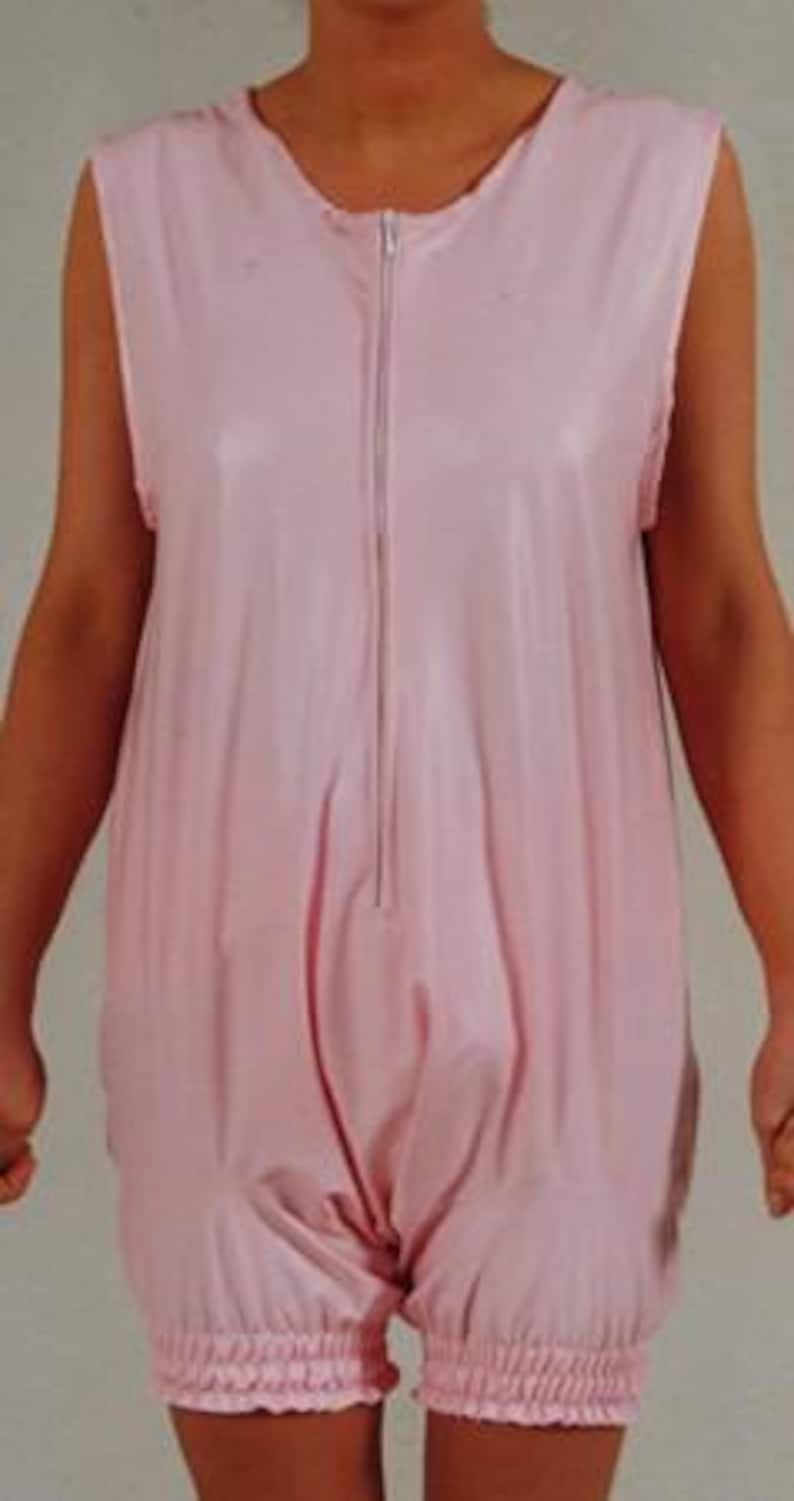 May include: A sleeveless, light pink romper with a front zipper. The garment has a gathered elastic hem at the shorts. The fabric appears to be a smooth, possibly waterproof material. The romper is designed with a loose fit.