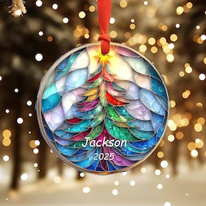 May include: A round Christmas ornament with a colorful stained-glass design of a Christmas tree. The ornament has the name "Jackson" and the year "2025" printed on it. It is suspended by a red ribbon.
