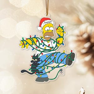 Op de afbeelding: Een heldere acryl kerstversiering met Homer Simpson verstrikt in kleurrijke kerstverlichting, met een zwarte kat. Homer draagt een kerstmuts met de naam "James". De versiering hangt aan een gouden draad, ideaal voor de feestdagen.