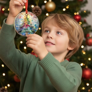 May include: A round Christmas ornament with a colorful stained-glass design of a Christmas tree. The ornament has a gold-colored border and a red ribbon for hanging. The name "Jackson" and the year "2025" are printed on the ornament.