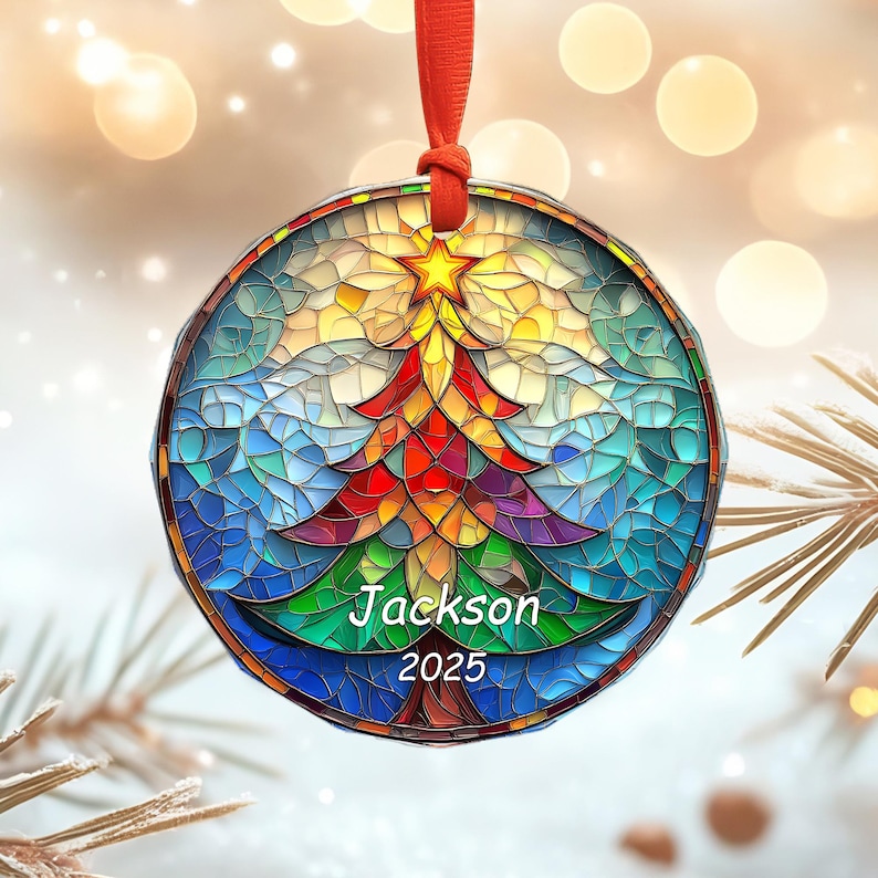 May include: A round Christmas ornament with a stained-glass design. The ornament features a colorful Christmas tree with a star on top, and the name "Jackson" and the year "2025" are printed on the tree. The ornament hangs from a red ribbon.