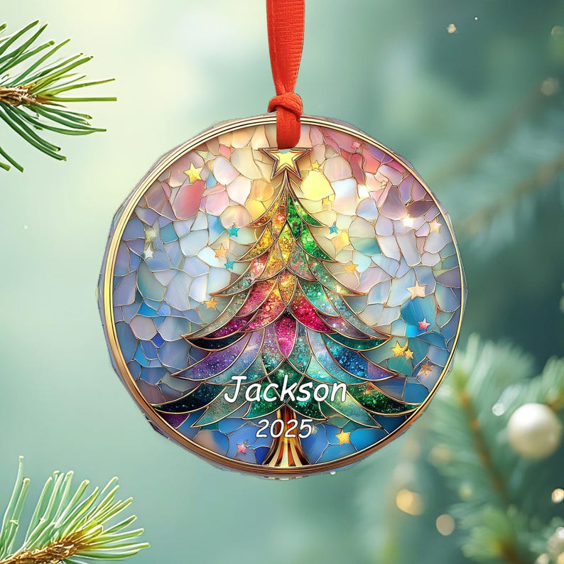 May include: A round Christmas ornament with a colorful stained-glass design. The ornament features a Christmas tree with the name "Jackson" and the year "2025". It hangs from a red ribbon. The ornament has a gold-colored border and is decorated with stars.