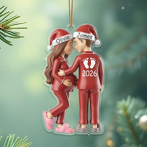 Può includere: Un ornamento natalizio raffigurante una coppia in pigiama rosso e cappelli di Babbo Natale. La donna è incinta e l'uomo la abbraccia. L'ornamento riporta i nomi "Olivia" e "James" e l'anno "2026".