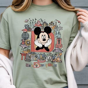 以下が含まれることがあります： セージグリーンのTシャツ。大きな「D」の中にミッキーマウスのカラフルなグラフィックがあり、ディズニーワールドのランドマークが描かれています。「Walt Disney World」の文字がデザインの下部にプリントされています。