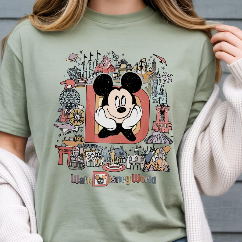以下が含まれることがあります： セージグリーンのTシャツ。大きな「D」の中にミッキーマウスのカラフルなグラフィックがあり、ディズニーワールドのランドマークが描かれています。「Walt Disney World」の文字がデザインの下部にプリントされています。