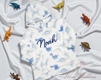 Pode incluir: Um moletom com capuz branco com estampa de dinossauros azuis e um bolso frontal. O nome Noah está bordado em letra cursiva azul-escura no peito. A peça está rodeada por várias pequenas figuras de dinossauros de plástico.