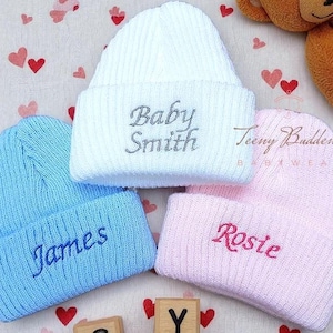 Pode incluir: Tr&ecirc;s gorros de beb&eacute; de malha em branco, azul e rosa. O gorro branco tem "Baby Smith" bordado em prata. O gorro azul diz "James" em azul, e o gorro rosa diz "Rosie" em rosa.