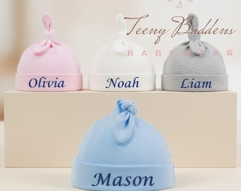 Pode incluir: Quatro chapéus de bebê em rosa, branco, cinza e azul, cada um com um top amarrado. Os chapéus são rotulados com os nomes "Olivia", "Noah", "Liam" e "Mason" em escrita azul marinho. Os chapéus são exibidos em um fundo neutro.
