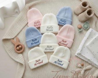 Pode incluir: Vários gorros de bebé em tons pastel, rosa, azul e branco, bordados com as frases "Hello World" e "Just Arrived". A imagem também apresenta botinhas de bebé, uma chupeta e um brinquedo de madeira.