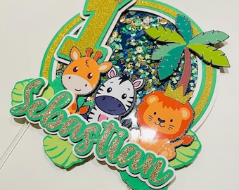 May include: A colorful cake topper featuring a giraffe, zebra, and lion with crowns. The number 1 and the name "Sebastian" are also included. The topper has a jungle theme with green and gold accents, and a palm tree.
