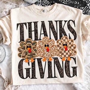 Puede incluir: Una camiseta color crema con el texto negro THANKS GIVING con textura agrietada. Entre las palabras hay tres pavos con picos y patas naranjas, cada uno con un patrón diferente en sus plumas: camuflaje, estampado de leopardo y cuadros.