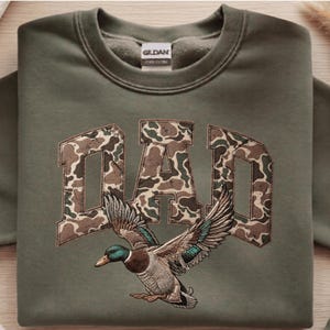 May include: An olive green sweatshirt featuring the word "DAD" in a camouflage design. Below the word is an embroidered duck with outstretched wings, in shades of green, brown, and blue.