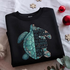 May include: Black sweatshirt featuring a teal sea turtle design. The turtle is split, with the left side detailed and the right side forming the words "AND INTO THE OCEAN I GO TO LOSE MY MIND AND FIND MY SOUL."