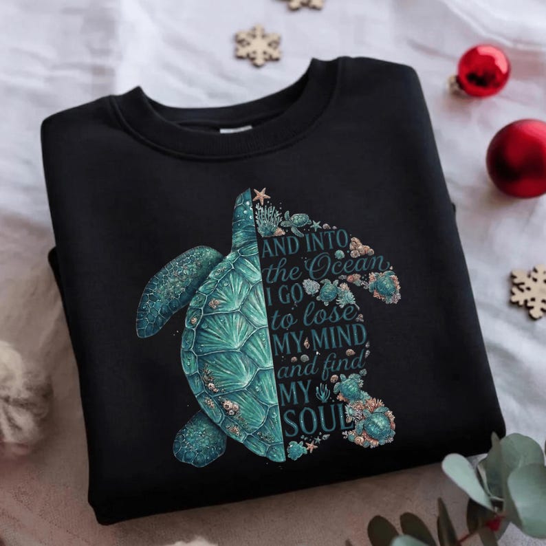 May include: Black sweatshirt featuring a teal sea turtle design. The turtle is split, with the left side detailed and the right side forming the words "AND INTO THE OCEAN I GO TO LOSE MY MIND AND FIND MY SOUL."