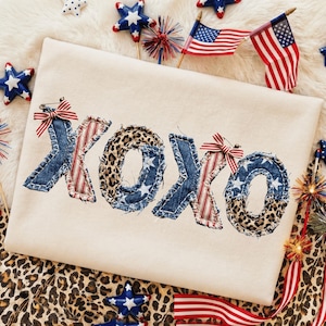 Op de afbeelding: Crèmekleurig T-shirt met het woord "XOXO" in een patchwork-ontwerp. De letters bevatten denim, luipaardprint en rood-wit gestreepte stof, met steraccenten. Patriotisch thema met Amerikaanse vlaggen en sterdecoraties.