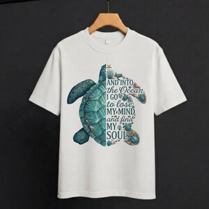 May include: White t-shirt featuring a detailed illustration of a sea turtle in shades of teal and green. The text "AND INTO THE OCEAN I GO to lose MY MIND and find MY SOUL" is printed on the right side of the turtle.