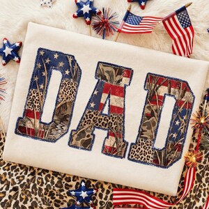 Può includere: T-shirt color crema con la scritta "DAD" in un design patchwork. Le lettere presentano motivi a bandiera americana, mimetici e animalier, bordati di blu. Decorazioni patriottiche circondano la maglietta.