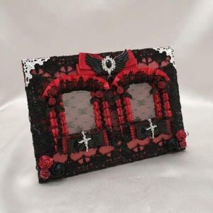 Puede incluir: Un marco de fotos rectangular con encaje negro sobre un fondo rojo. Incluye dos aberturas arqueadas, volantes de cinta roja, lazos negros, pequeñas rosas de tela rojas y negras, detalles plateados en las esquinas y un emblema central de alas negras.