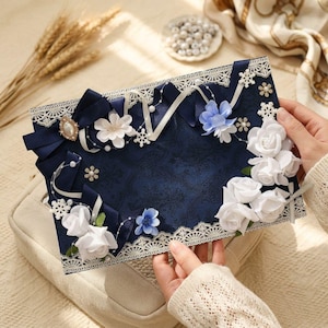 Könnte beinhalten: Eine rechteckige marineblaue Karte mit dunklem Damastmuster, verziert mit weißer Spitzenborte, weißen und blauen Stoffrosen, marineblauen Bändern, kleinen Perlen und weißen Schneeflocken-Akzenten. Die Karte hat eine ovale Brosche in der Mitte.