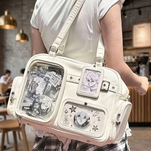 May include: A white ita bag with a clear front pocket displaying anime-style character illustrations, circular pin-back buttons, and star-shaped charms. The bag features a vertical rectangular card holder with a character print and side pockets.