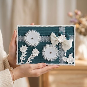 Puede incluir: Un tablero rectangular de pana verde azulado oscuro con un borde de encaje blanco y una tira de encaje horizontal. Está decorado con dos almohadillas de fieltro circulares, un lazo de tela blanco con una margarita y apliques florales.