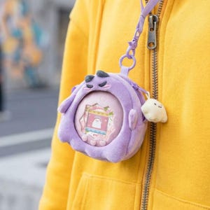 May include: A purple plush protective case for a digital pet device, featuring a rounded shape with embroidered facial features. The case includes a purple strap with a metal clip and a small, cream-coloured plush charm attached to the side zipper.